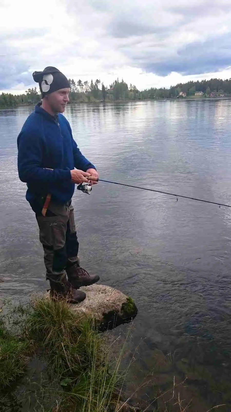 En person fiskar vid sjön på Kvissleströmmens fiskecamping, omgiven av naturskön miljö, med ett fiskespö i handen och fokus på det stilla vattnet.