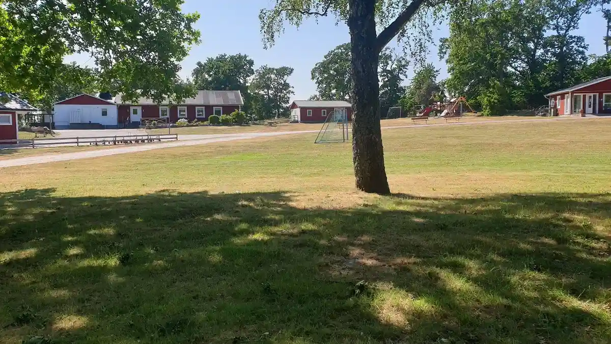 En somrig bild visar Kustgården Senoren, med en blå himmel och gröna träd. I förgrunden finns gräs och en öppen yta, medan ett hus och bostadsområde syns i bakgrunden.