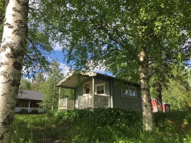 En idyllisk bild av Norråkers camping som visar en lantlig stuga omgiven av grönskande träd och buskar. I bakgrunden syns en tät skog, vilket förstärker den fridfulla och naturnära atmosfären.