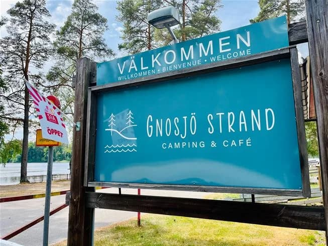 Alt-text: En skuggig trädgård med utomhusmöbler och ett bord, omgiven av gröna växter och byggnader i bakgrunden vid Gnosjö strand.