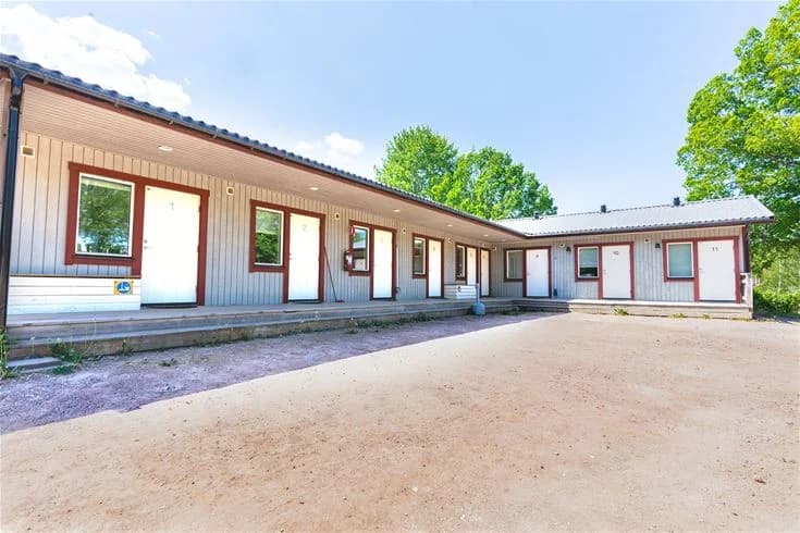 En liten stuga vid Vimmerby Camping - Nossenbaden, omgiven av en gräsmatta och en enkel uppfart. Det är en kylig dag och stugans ytterväggar är klädda med träpanel.