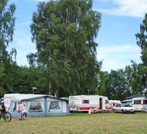 En grön campingplats med träd, husbilar, tält och en husvagn, omgiven av frodig gräsmatta och parkeringsyta på Bödagårdens camping.