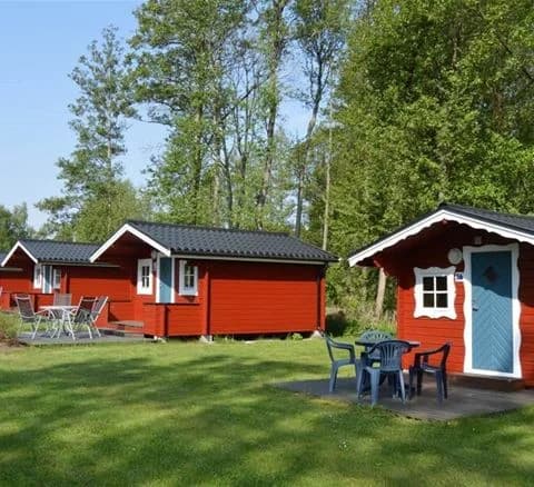 Bilden visar en stuga omgiven av en välskött gräsmatta och trädgård på Bödagårdens camping. Det finns utemöbler och trädgårdsbyggnader, vilket skapar en inbjudande atmosfär för besökare.