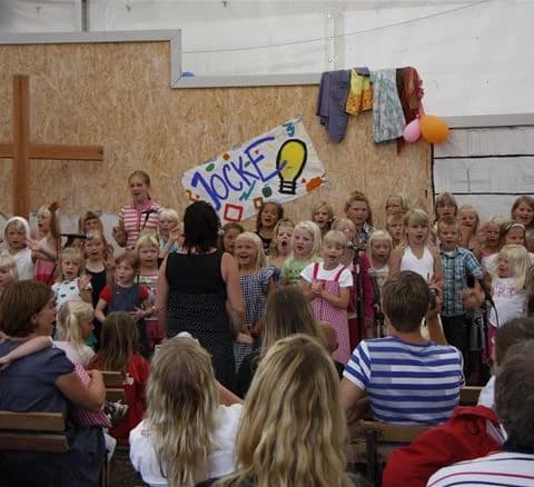 En grupp människor, inklusive barn och ungdomar i T-shirts, deltar i en utomhusaktivitet på Bödagårdens camping. Stämningen är glad och avslappnad med fokus på gemenskap och underhållning.