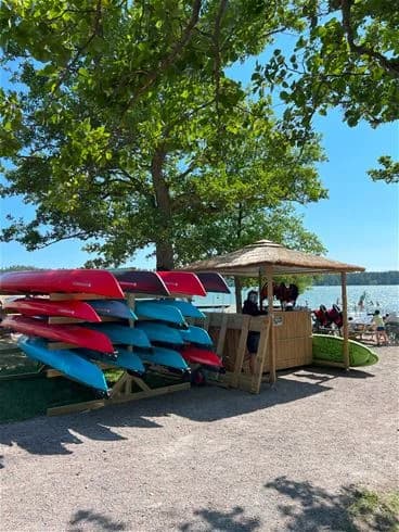 En solig sommardag vid Trosa Havsbad & Camping, med båtar som ligger förtöjda vid kusten och färgglada parasoller som ger skugga åt besökare på stranden. Människor njuter av friluftsliv och avslappning nära vattnet.