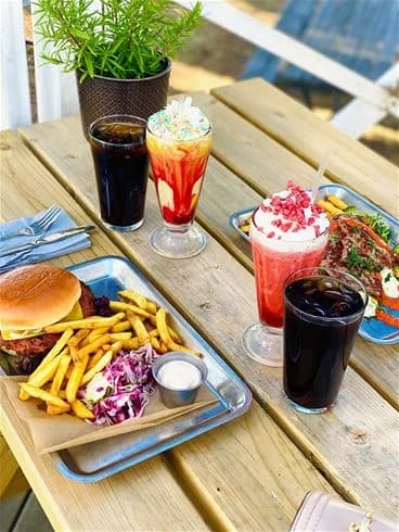 En bild som visar ett bord uppdukat med snabbmat. Det finns pommes frites och andra friterade maträtter, tillsammans med drycker serverade med sugrör. Bordet är en del av serveringen vid Trosa Havsbad & Camping.