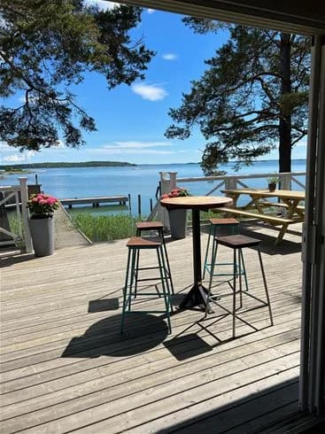 En träbord och stolar står på en trädeck vid Trosa havsbad & camping, omgivna av skugga och naturlig, träfärgad inredning.