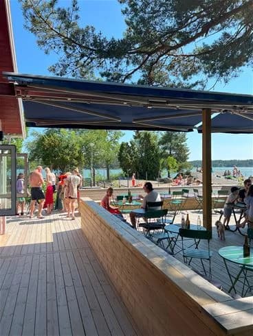 Bild av Trosa Havsbad & Camping visar ett soligt fritidsområde med utomhusmöbler under skugga. I bakgrunden syns en brygga och en gångväg vid vattnet, vilket förstärker semesterkänslan i den pittoreska badorten.