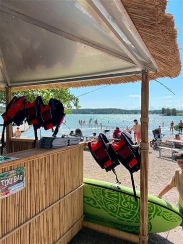 En solig dag på Trosa Havsbad & Camping med människor som njuter på stranden i sina badkläder. Stranden är täckt av sand och skuggas delvis av parasoller, vilket skapar en avslappnad atmosfär för semesterfirare.