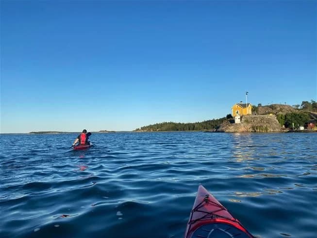 En grupp människor paddlar i kajaker på lugnt vatten nära en kustlinje vid Trosa Havsbad & Camping. I bakgrunden syns några fritidsbåtar och skogsklädda landformer.