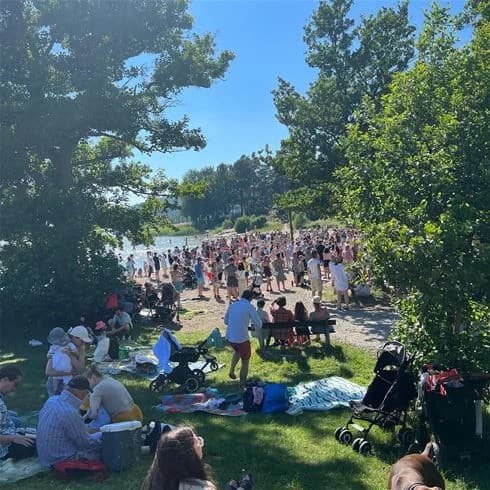 En stor grupp människor som njuter av en solig dag på Trosa Havsbad & Camping, där de deltar i olika sommaraktiviteter och avkopplande stunder vid stranden.