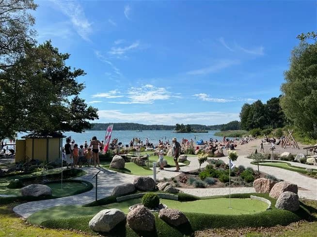 En solig dag vid Trosa Havsbad & Camping med människor som njuter av fritidsaktiviteter och picknick. Platsen är omgiven av tropisk grönska och utrustad med utomhusmöbler för avkoppling och semesterfirande.