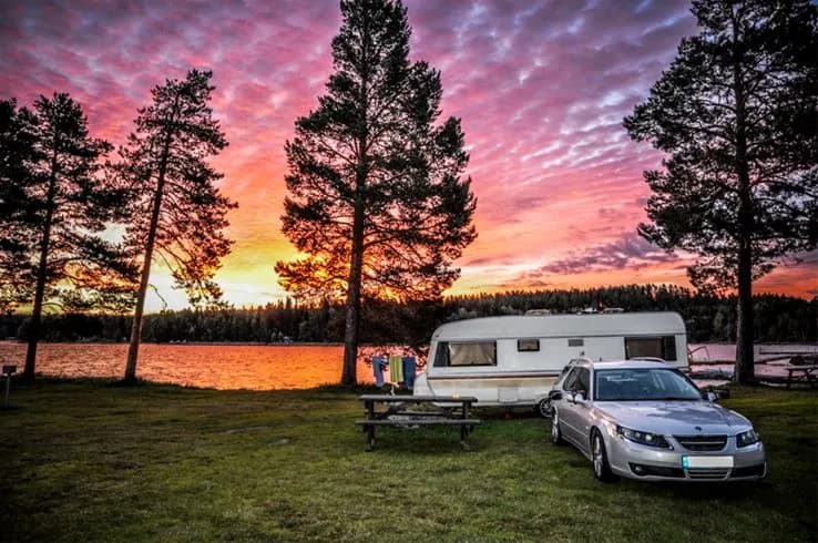En husbil står parkerad vid en sjö i solnedgången, omgiven av mjukt efterglödsljus och spegelreflektioner i vattnet, vilket skapar en rofylld stämning vid Camp Viking.