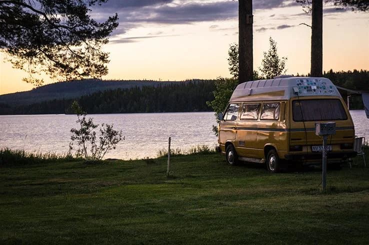 En vit Volkswagen minibuss står parkerad vid ett naturskönt område med skogar och en sjö i bakgrunden, vilket ger intrycket av ett fridfullt resmål.