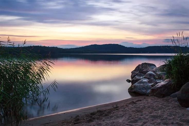 En vacker vy vid Skotteksgården Camping & Stugby, med ett stilla vatten som speglas i skymningen, omgiven av stora stenar och en blå himmel vid horisonten.