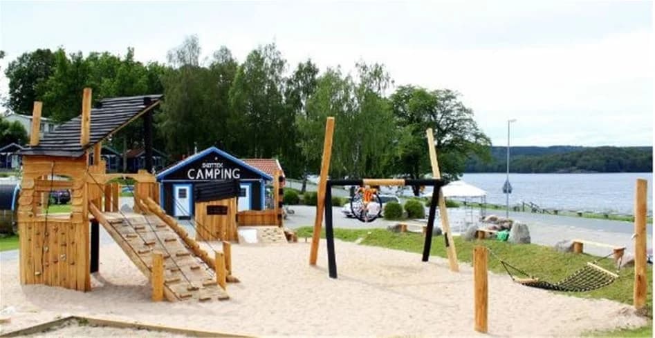 En campingplats med lekutrustning bland träd som ger skugga. Sand och utemöbler syns, vilket skapar en avslappnad miljö för fritidsaktiviteter.