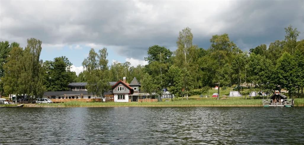 En fridfull scen vid Vimmerby Camping - Nossenbaden med en liten båt som flyter på en stilla sjö, omgiven av träd längs strandkanten.