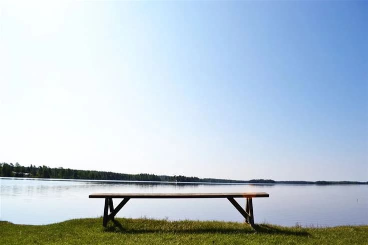 En somrig vy över Gökaskratts camping med en klarblå himmel och horisont, intill en sjö omgiven av grönska. En utomhusbänk står vid sjön och inbjuder till avkoppling och njutning av naturen.