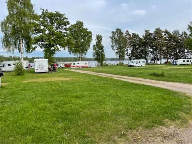 En campingplats med husvagnar och husbilar parkerade på gräsytor under en blå himmel med moln. I bakgrunden syns ängar och åkrar, vilket ger ett naturskönt intryck.