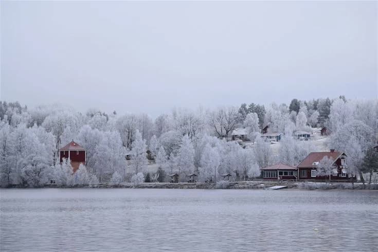 En vinterbild av Sandaholm Restaurang & Camping. Huset är täckt av snö och omgivningen är frusen, vilket ger en mysig atmosfär. Grenar är delvis begravda i snön och platsen liknar en idyllisk vinterstuga i en liten by.