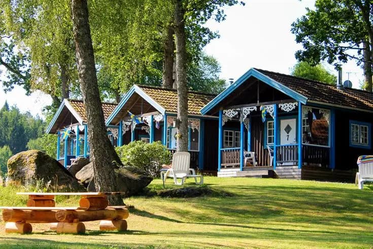 En stuga omgiven av en grönskande trädgård med gräsmatta och skuggiga områden på Skotteksgården Camping & Stugby. En veranda och andra utomhusstrukturer syns i bilden, vilket ger en känsla av avkoppling och friluftsliv.
