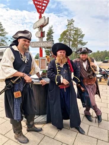 En person klädd som pirat, med solhatt och skägg, står vid Stenö havsbad & camping. I bakgrunden syns campingområdet och blå himmel.