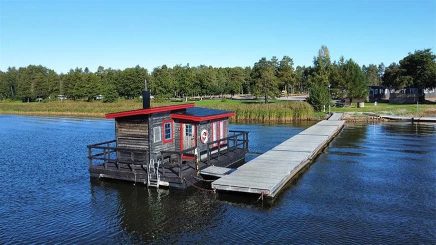 Vy över Stenö havsbad & camping med en lång brygga som sträcker sig ut över en lugn vattenyta, omgiven av grönska vid strandkanten.