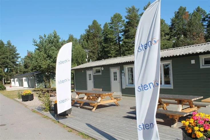 En campingplats vid Stenö Havsbad med utomhusmöbler på en gräsmatta och en byggnad med dörr och skugga i bakgrunden.