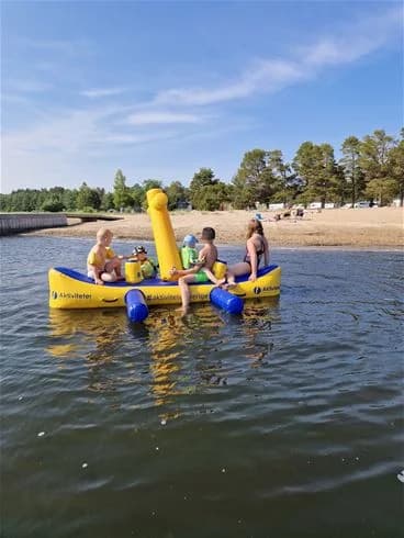 En grupp människor njuter av utomhusaktiviteter på Stenö Havsbad & Camping. Flera uppblåsbara båtar och vattenleksaker ses på vattnet, medan besökare deltar i fritidsaktiviteter och rekreation på den natursköna campingplatsen.