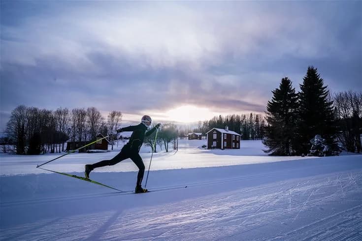En person åker längdskidor på snötäckta spår, omgiven av vinterlandskap. I bakgrunden syns fler skidåkare och träd klädda i snö. En perfekt dag för vintersport på Åsarna Skicenter.