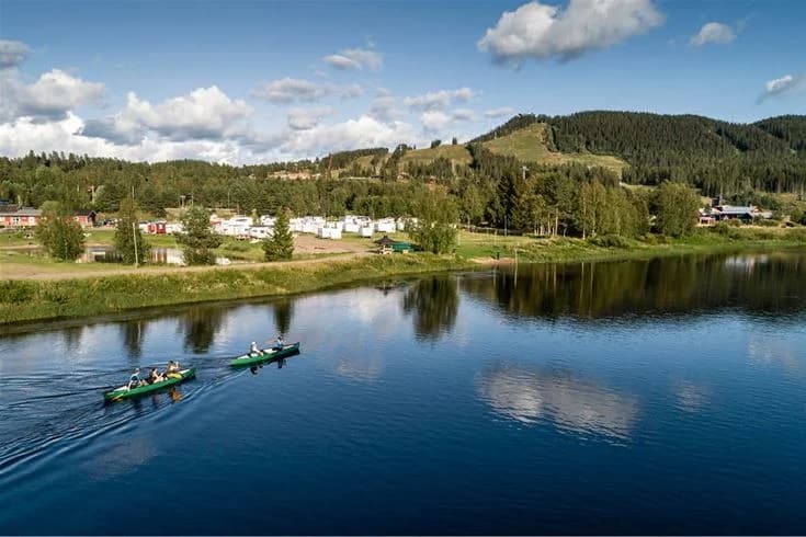 En somrig landskapsbild av Kläppens camping med grönska och träd som omger en vattenväg. Solbelysta vattenreflektioner syns på ytan vid strandkanten.