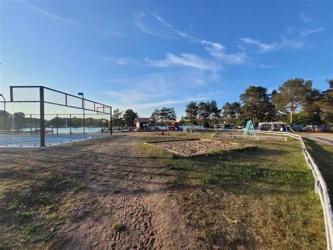 En solig dag vid Älvkarleby fiskecamping med moln på himlen, omgiven av grönska och ett staket i förgrunden.