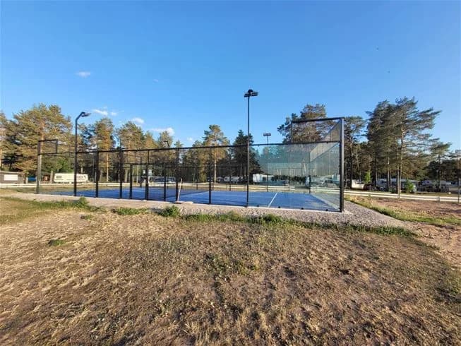 Bild av en solbelyst sportanläggning vid Älvkarleby fiskecamping med skuggor från stängslet runt platsen, som påminner om ett tennisområde. Ett stängsel av nät omger platsen.