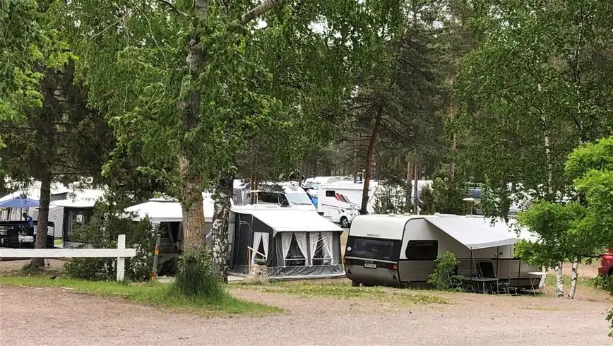 En fridfull campingplats i Älvkarleby med husvagnar och tält uppställda under skuggande träd. Flera fordon är parkerade i området, vilket ger en inbjudande atmosfär för rekreation och avkoppling i naturen.