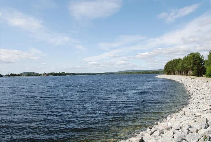 En blå himmel sträcker sig över en lugn vattenyta vid Storumans camping, omgiven av moln och en horisontlinje som markerar en vattenväg och kustlandskap.