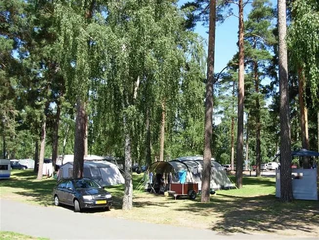 En campingplats med träd och gröna växter som ger skugga. Det finns ett tält uppsatt och bilar parkerade i närheten, omgiven av ett parkliknande område.