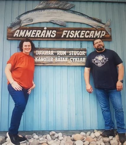 En glad man med skägg och mustasch håller upp en stor fisk vid Ammeråns fiskecamp. Han bär en skjorta med en logo och ler mot kameran.