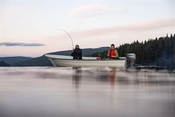 En liten motorbåt ligger förtöjd vid sjöns strandkant vid Ammeråns fiskecamp, med spegelblanka vattenytor som reflekterar himlen och omgivande natur.