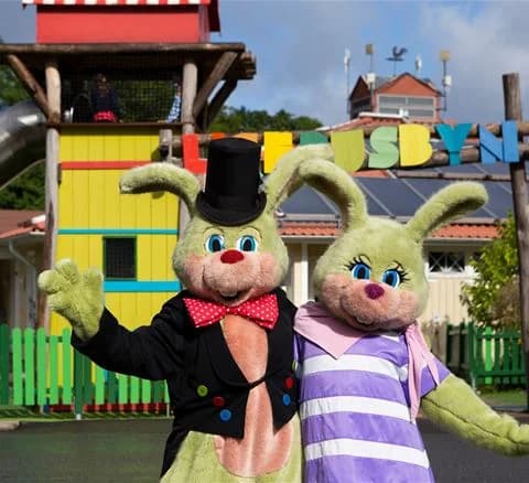 En glad maskot iklädd kostym ler och vinkar, omgiven av glada besökare som njuter av roliga aktiviteter och fritid på Lisebergsbyn.