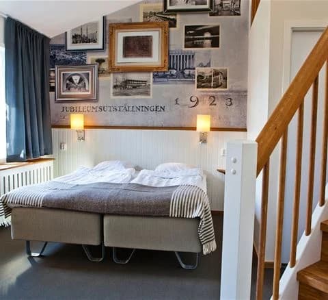 Ett stilrent sovrum på Lisebergsbyn med en stor, inbjudande säng, vit sängklädsel och smakfull inredning. Väggarna pryds av en enkel tavelram, och trädetaljer syns i både golvet och möblerna.