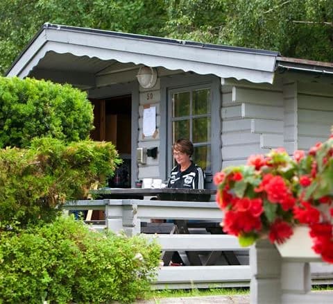 Bild av Lisebergsbyn med buskar, en stuga och en trädgård med utemöbler. Gräsmatta och trädgårdsbyggnader syns i bakgrunden, vilket skapar en avkopplande atmosfär.