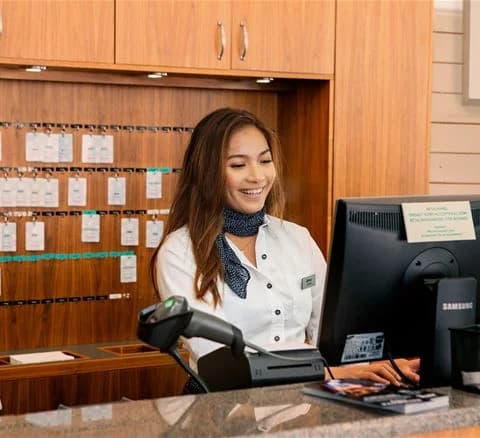 En receptionist i Lisebergsbyns välkomnande kontor, leende vid disken med en dator och en telefon nära till hands, redo att hjälpa besökare.