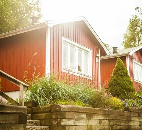 Alt-text: En idyllisk stuga omgiven av gröna buskar och välskött trädgårdsmiljö, symboliserande vårkänsla i Lisebergsbyn.