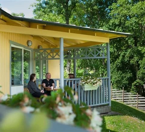 En mysig stuga på Lisebergsbyn med en veranda som erbjuder skugga, omgiven av en välskött trädgård med gräsmatta och trädgårdsbyggnader i bakgrunden.