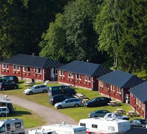 En bild av Lisebergsbyn som visar en parkering med flera bilar, omgivna av gröna träd och en välskött gård.