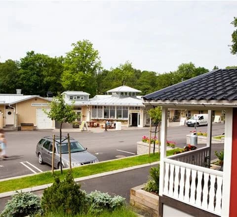 En idyllisk vy av Lisebergsbyn med flera röda stugor som omges av grönskande träd och välskötta gräsmattor. Stugorna har vita dörrar och fönsterluckor, och mellan dem slingrar sig en asfalterad gångväg. I bakgrunden syns en skog som förstärker naturkänslan.