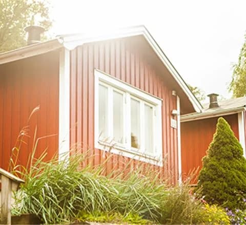 En röd stuga med vita knutar omgiven av grönskande buskar och trädgård, vilket ger en inbjudande och hemtrevlig känsla vid Lisebergsbyn.