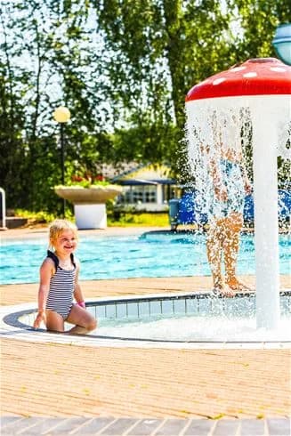 En somrig dag på Harge Bad & Camping där barn i badkläder leker och har roligt vid en utomhuspool. Atmosfären är avslappnad och inbjuder till lek och avkoppling under semestern.