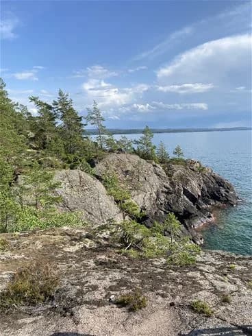 Vy över Harge Bad & Camping, med en karg kustlinje som kantas av steniga klippor. Vattnet från sjön breder ut sig i bakgrunden, och längs kusten syns den natursköna terrängen som består av klippor och stränder. Sommarsolen lyser över det vilda landskapet och skapar en idyllisk sommarstämning.