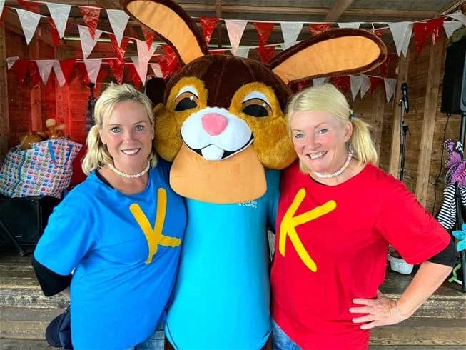 En grupp glada människor, inklusive barn, klädda i lekfulla dräkter framför en maskot på Harge Bad & Camping. De ser ut att njuta av en solig dag fylld med lek och avkoppling.